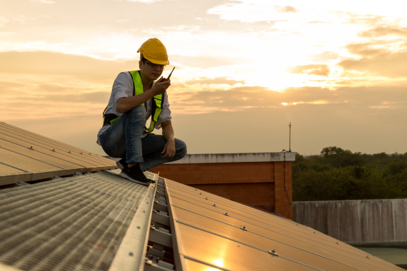 A solar technician in a hard hat and safety vest sitting on a commercial rooftop next to installed solar panels during a site assessment.