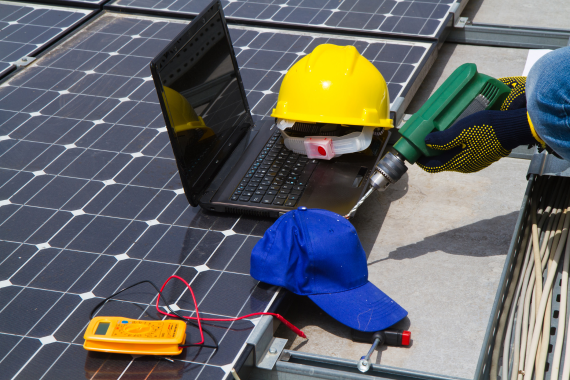 Close-up of a laptop, multimeter, and hard hat on a solar panel during the installation and testing phase.