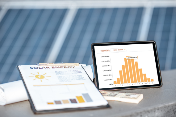 A tablet and a printed solar report showing energy data and an orange bar chart, lying next to a solar panel.