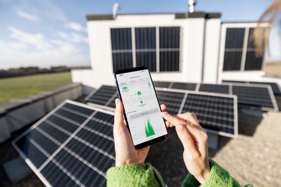 A hand holding a smartphone displaying a solar monitoring app in the foreground of an installed solar panel array.