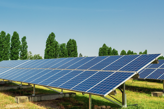 Rows of ground-mounted utility-scale solar panels in a field under a bright blue sky.