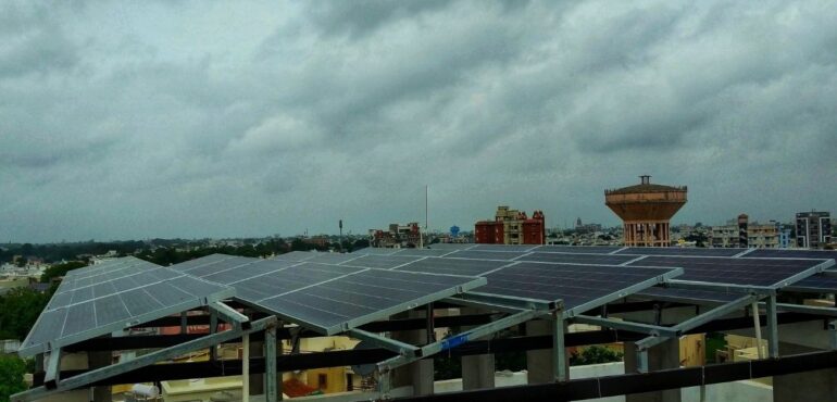 A large array of solar panels mounted on a building rooftop in an urban area under a cloudy sky.