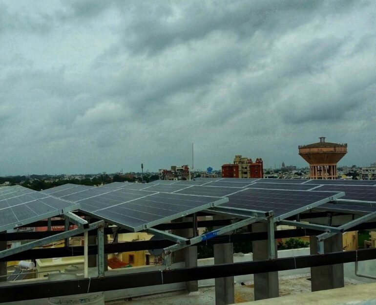 A large array of solar panels mounted on a building rooftop in an urban area under a cloudy sky.