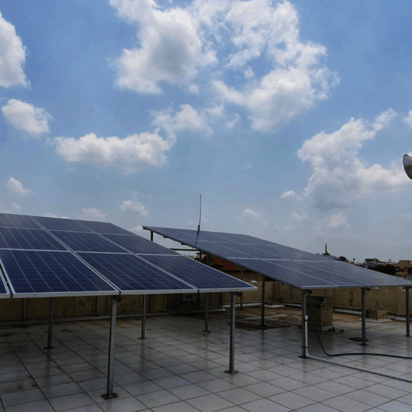 Solar panels installed on an elevated mount on a flat rooftop against a bright blue, cloudy sky in Nadiad.