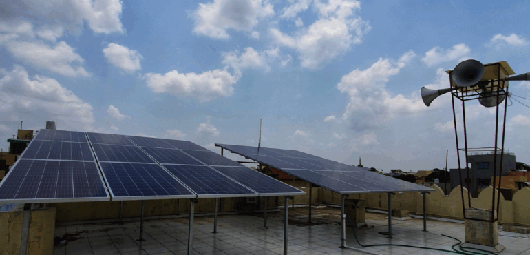 Solar panels installed on an elevated mount on a flat rooftop against a bright blue, cloudy sky in Nadiad.
