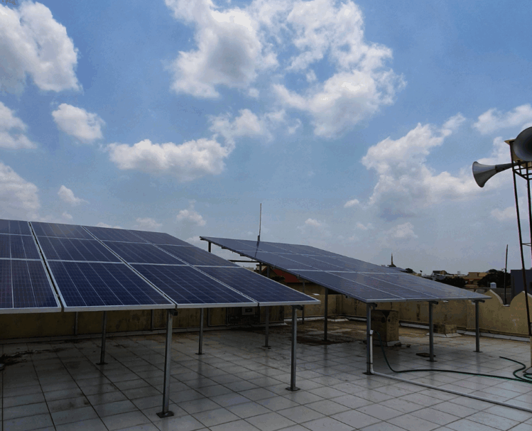 Solar panels installed on an elevated mount on a flat rooftop against a bright blue, cloudy sky in Nadiad.