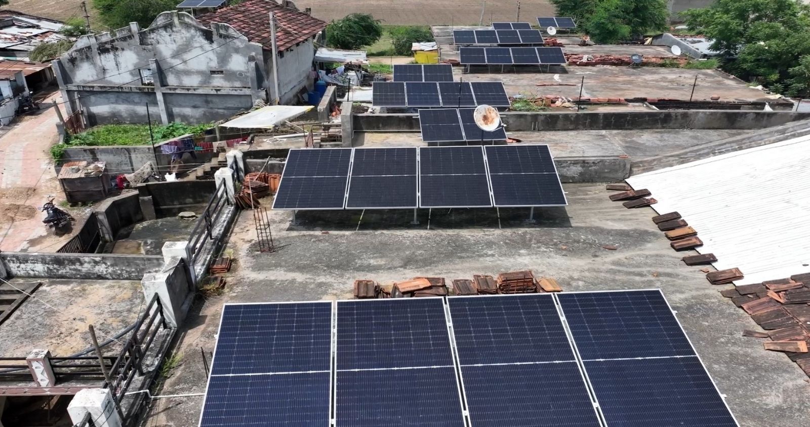 A wide view of multiple solar panel arrays installed on the flat rooftop of a building in a village setting.